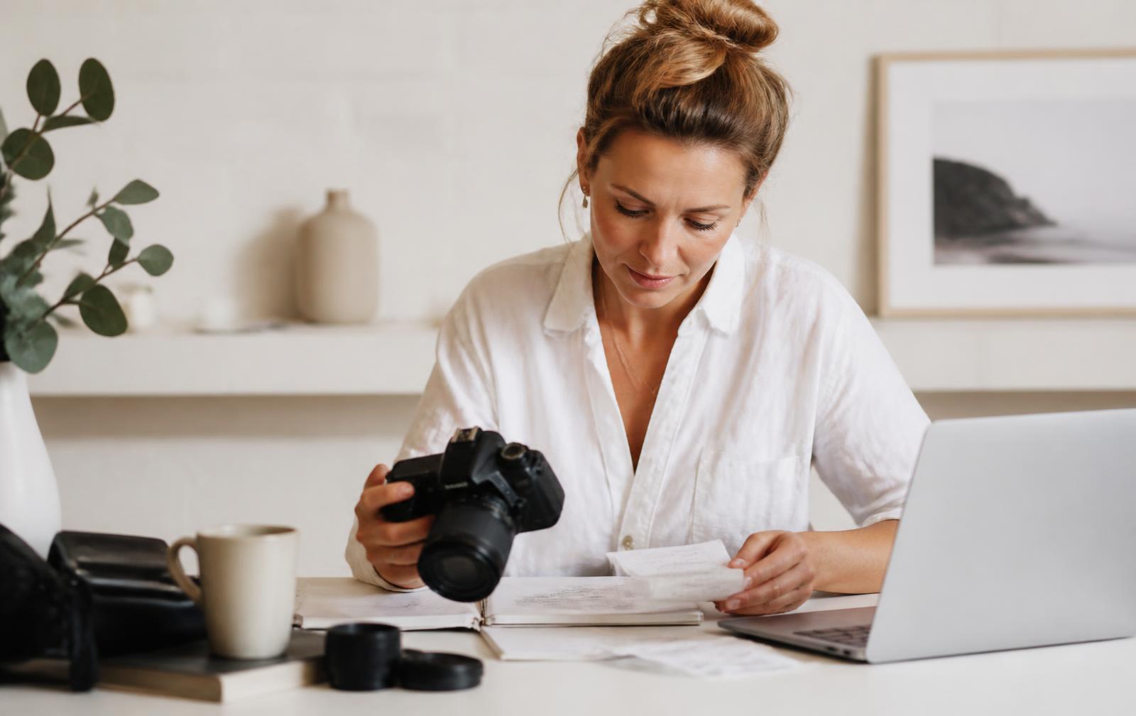 Freelance photographer reviewing receipts at a sunlit desk with a camera and laptop