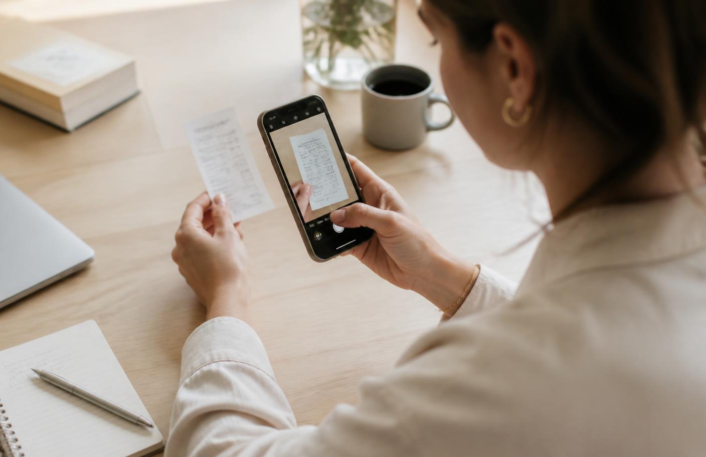Sole trader photographing a paper receipt with a smartphone at a light wooden desk
